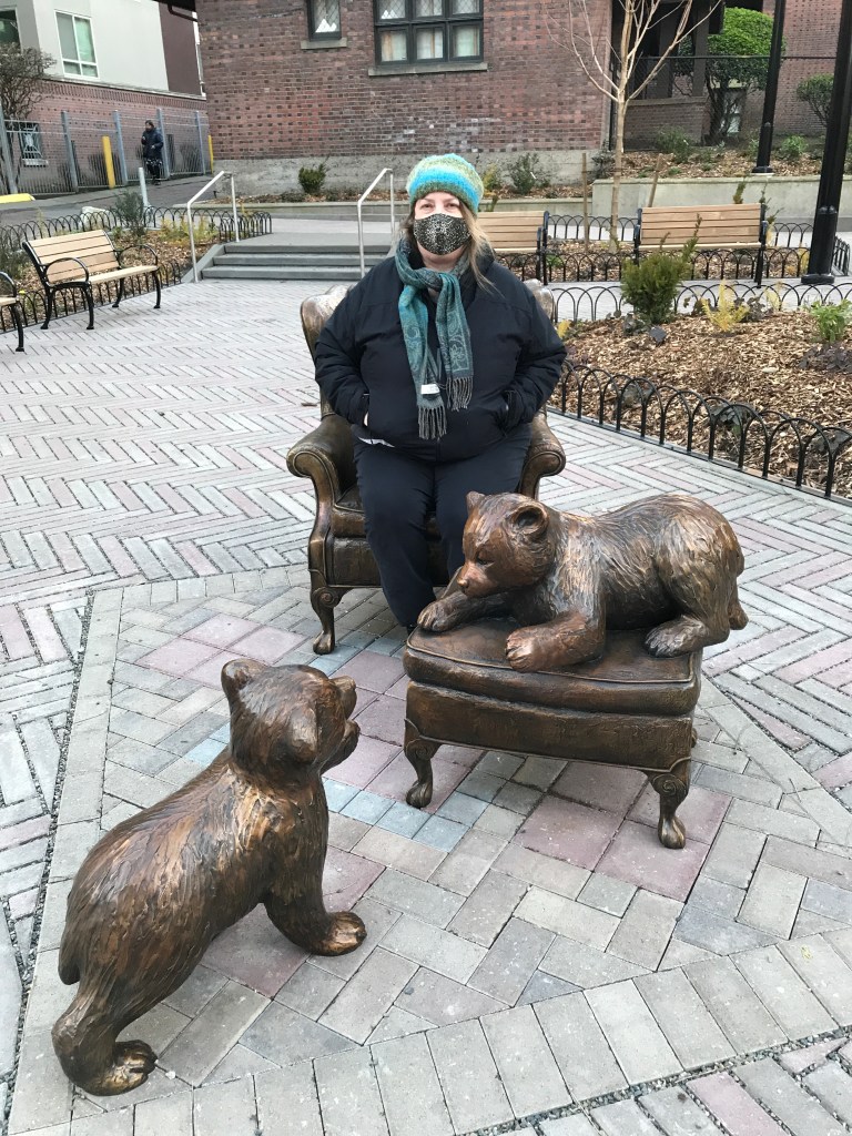 Women with mask sitting in bronze chair of statue with bronze bear cubs in small park. 