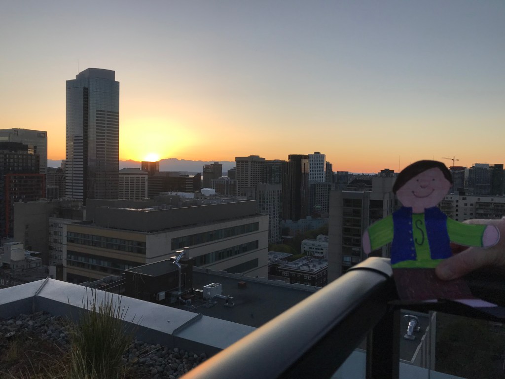 Paper cut out of boy on high rise roof deck with city and sunset behind him. 