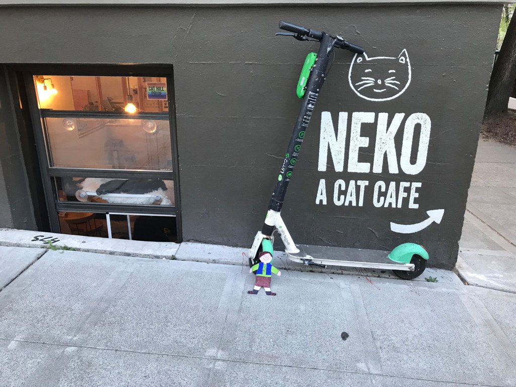 Paper cut out of boy in front of scooter next to a sign reading "Neko A Cat Cafe" with cat lounging in window. 