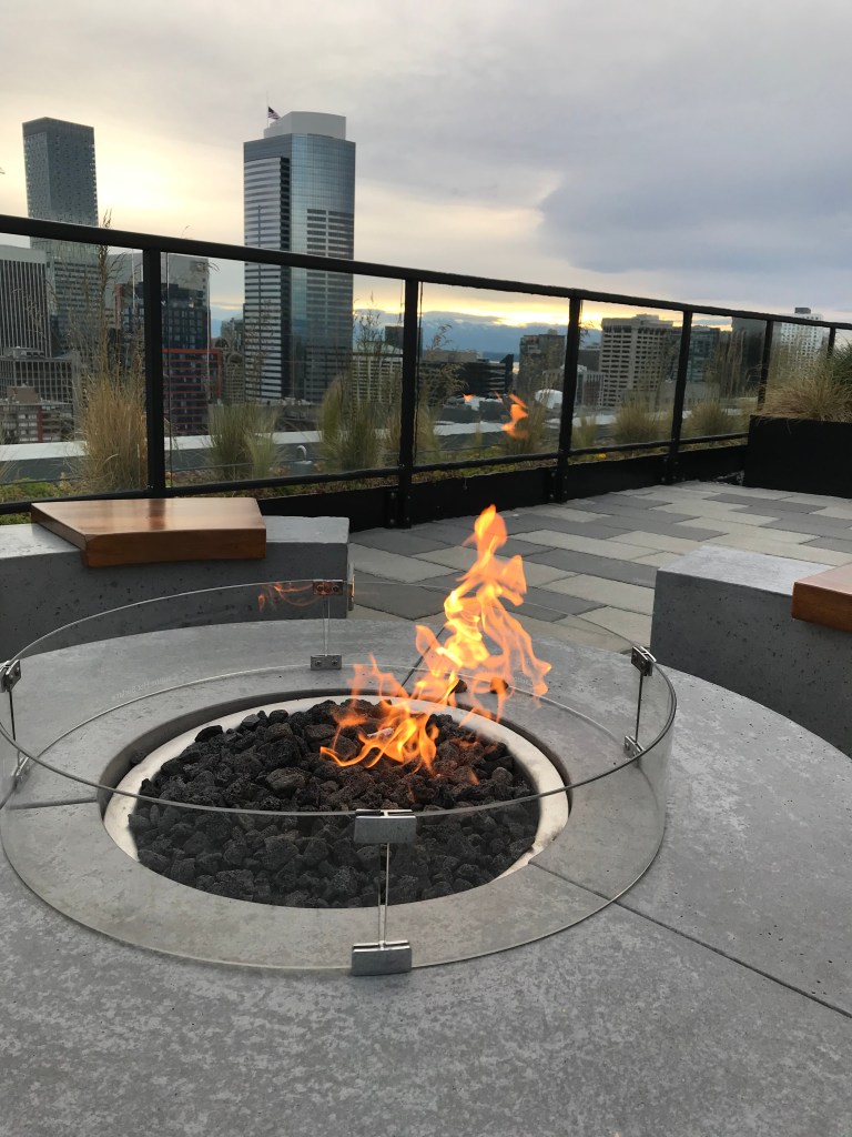Fire in circular fire pit on roof of tall building, other skyscrapers in the background. 